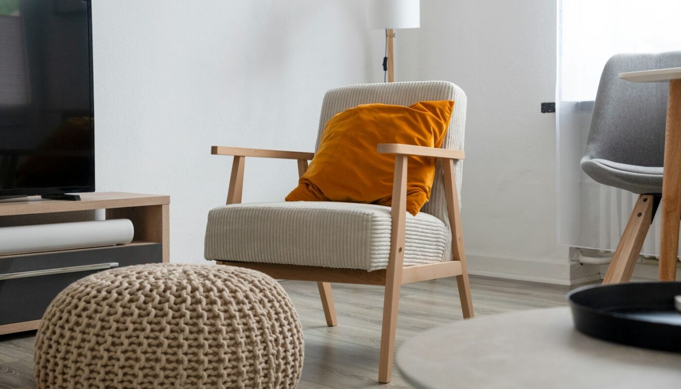 A living room with a wooden armchair, orange cushion and knitted pouffe (photo: eskoala / Unsplash)