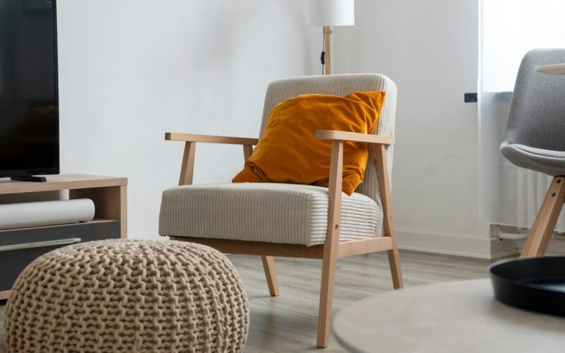 A living room with a wooden armchair, orange cushion and knitted pouffe (photo: eskoala / Unsplash)