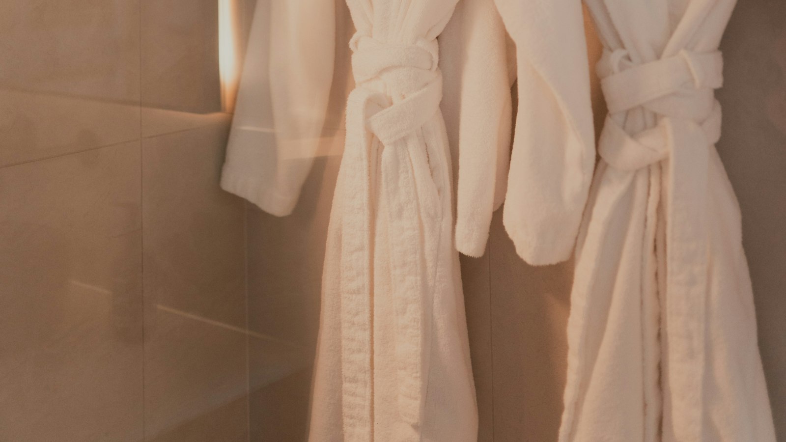 Two white bathrobes hanging on hooks in a warm-lit modern bathroom (photo: Grace Anne Bobadilla / Unsplash)