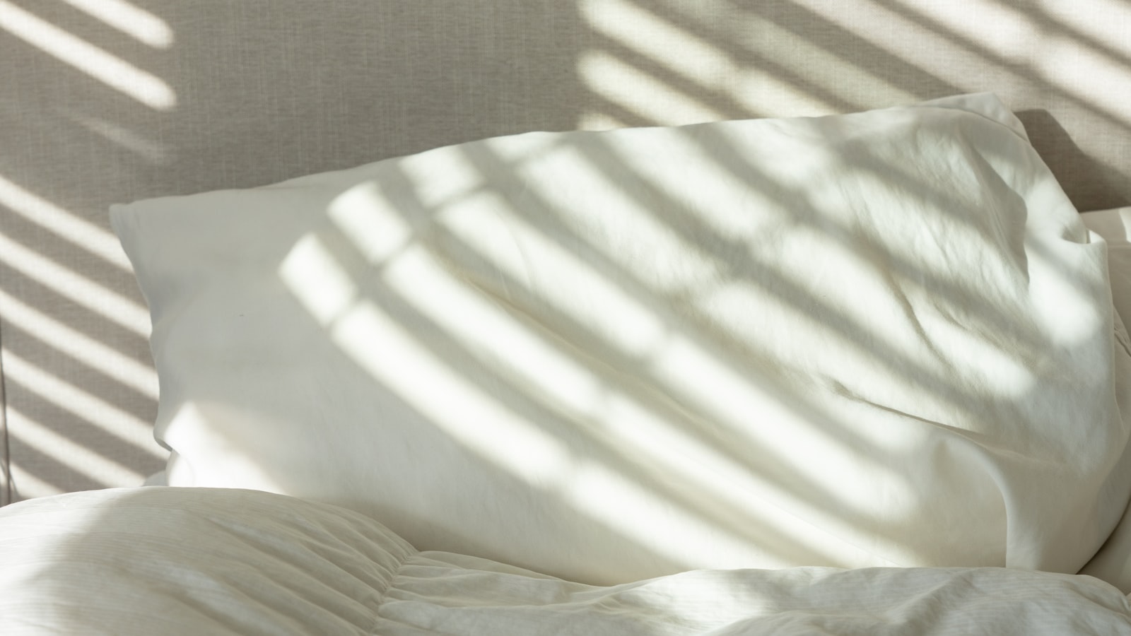 A white pillow on a made bed with striped morning sunlight falling across it (photo: Liz Vo / Unsplash)